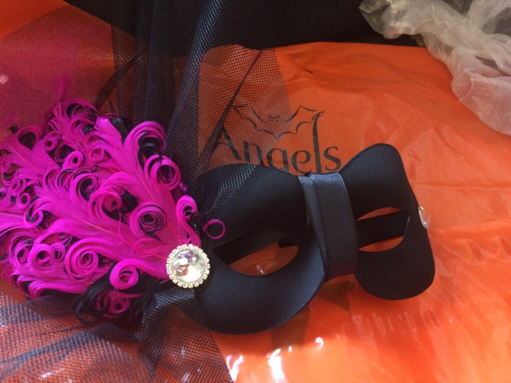 A black masquerade mask on top of an orange shopping bag, with fuchsia feathers and a glittering plastic jewel holding them on. 