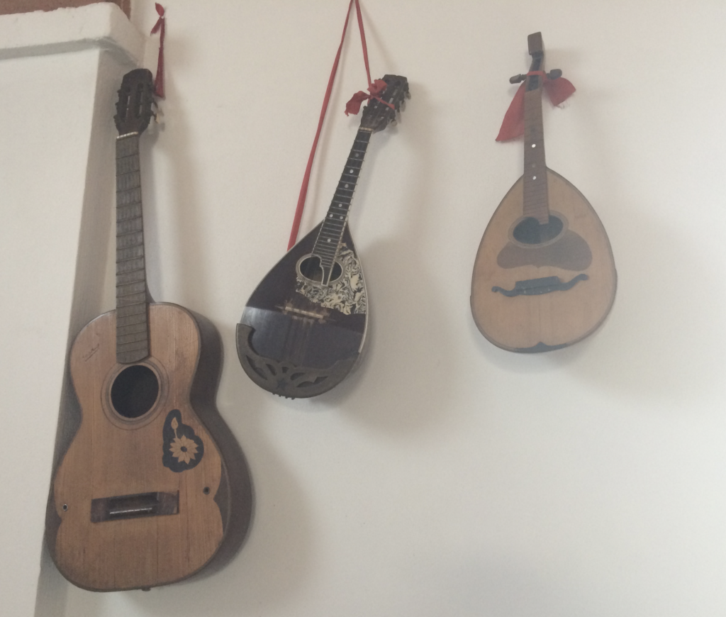 Three stringed instruments hang from a wall. The centre instrument is askew. The left-side one is a more modern guitar, but with details and coloring suggesting it is old. The other two are black and natural wood, respectively, and appear to be lutes. The right-side instrument has a bow tied around its neck. Its bridge looks like a mustache.