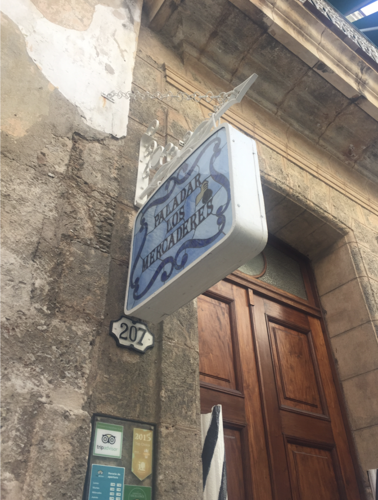 The image shows a wooden door in a weathered stone building with the number 207 to the left of the doorway and a sign stating "Paladar los Mecaderes" with blue, stained-glass style lettering. Below the number 207, there are a variety of small, unreadable posters and badges from TripAdvisor.