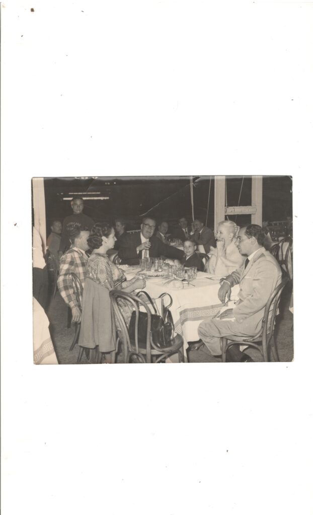 A black-and-white image of a group of people seated around a table. The man at the head of the table appears to be holding a drink.